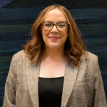 A colour portrait of Sophie Baillie stood against a blue-green background. Sophie is a white woman with shoulder length hair who is wearing a beige jacket and black top.