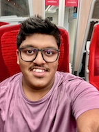 A portrait of Surya Jothi taking a selfie on a train with red seats. Surya is an Asian man with short dark hair, glasses and wearing a mauve t-shirt.