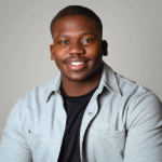 A colour portrait of Tariq Peters smiling at the camera. Tariq is a Black man with short cropped hair who wears a grey shirt over a black t-shirt. The background is grey