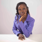 A colour portrait of Tofunmi Akinde. Tofunmi is a Black woman with long hair, wearing a purple jacket, with one had rested on her chin. The background is grey.