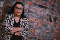 Advita Patel stood against a brick wall. Advita is an Asian woman with glasses, dark hair and wears a leopard print coat. Her arms are crossed