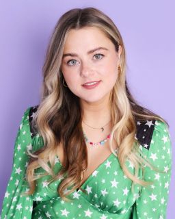 Ashleigh Skinner, a white woman with long blond hair, wears a green dress with white star patterns. The background is mauve. She smiles looking at the camera