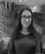 A portrait of a white woman with long dark hair with a dark top on and glasses