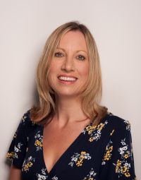 Carol Middleton, a white woman with shoulder length blond hair, smiles at the camera. She wears a navy patterned top. The background is grey