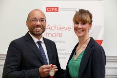 Cornelius Alexander, a black man with glasses and a short dark bear, stands in a suit and tie smiling. He holds a medal. To his right is Sarah Hall, a white woman with light brown hair, wearing a dark blazer and green top.