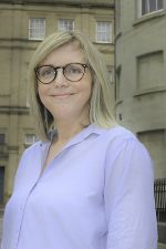 A smiling white woman with blond hair, glasses wearing a white shirt stands in front of a building