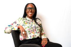 A smiling Elizabeth Banuka, a Black woman with glasses, wears a floral shirt while sat sideways and facing the camera