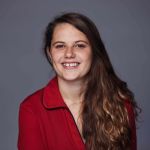 A portrait of a smiling Ella Percival against a grey backdrop. Ella is a white woman with long brown hair who wears a red top.