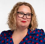 A portrait of Emma Duke wearing a red spotted blue blouse. Emma is a white woman with curly blonde hair and wears glasses