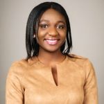 A portrait of Esenam Agubretru smiling against a grey background. Esenam is a Black woman with shoulder length dark hair and wears a gold blouse