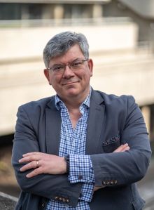 Guy Clapperton with his arms crossed. Guy is a white man with short grey hair who wears glasses, a grey blazer and blue check shirt. The background is blurred.