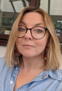 Kat Dibbits looking at the camera. Kat is a bite woman with shoulder length blond hair and glasses, wearing a blue shirt