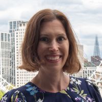 A smiling Kate Busby, a white woman with brown hair. She is stood in front of the London skyline