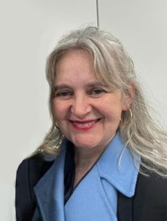 A portait of Lora Benson, a white woman with long grey hair, smiling at the camera. She wears a black jacket with light blue collar
