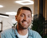 A white young man with cropped hair and stubble iis wearing a blue shirt with a white t-shirt underneath - an office light is above himand a plant behind him