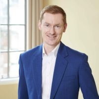Stuart Skinner, a white clean-shaven man with red hair, wearing a blue blazer and white open neck shirt. He stands in front of a window, cream curtain and cream wall