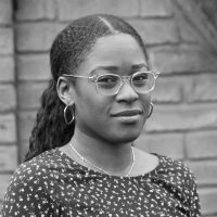 Black and white image of Tani Fatuga, a Black woman with a pony-tail, glasses and earrings. She wears patterned top. The background is a brick wall.