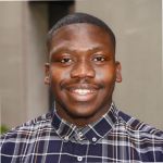 A portrait of Tariq Peters against a grey background. Tariq is a Black man with cropped hair, wearing a purple check shirt