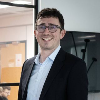 Thomas Averre, a white man with short dark hair and glasses, wears a dark jacket and light shirt, while smiling at the camera. Behind him is a screen and a window to an office where colleagues are sat