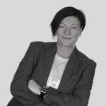 A black and white portrait of Gaby Jesson. Gaby is a white woman with dark hair, wearing a blazer, with her arms crossed.