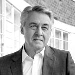 A black and white portrait of Jon White in front of a building. Jon is a white man with short hair, wearing a shirt and jacket.