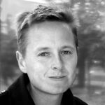 A black and white portrait of Christian Koch. Christian is a white man with short light hair. There are trees in the background.