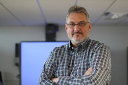 Alastair McCapra, a white man with grey hair and beard and wearing glasses, stands in a check shirt with his arms folded