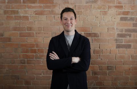 Andy McCaul with arms crossed and smiling at camera. Andy is white with dark hair, wearing a black jacket. He is pictured against a brick wall.