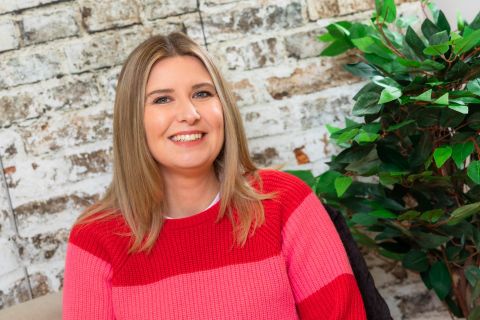 A photo of a smiling Kat Trinder looking straight to camera. She is wearing a red and pink horizontal striped top and has shoulder length blonde hair. Behind Kat is a brick wall and to her left is a large plant.