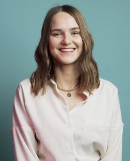 Claire Simpson smiling at the camera. Claire is white with brown shoulder length hair. Claire wears a necklace and pale pink shirt. The background is turquoise.