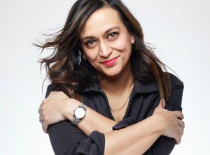 Shilpa Saul, a British Asian woman with dark shoulder length hair, red lipstick. She wears a black shirt with sleeves rolled up. Her arms are crossed reaching towards her shoulders. She wears a watch on her left hand