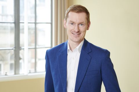 Stuart Skinner is a white man wearing a blue jacket and white shirt. He is looking at the camera and smiling. He is stood against a magnolia wall with a window on the left hand side of the photograph.