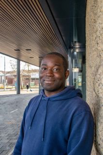 Ọmọ́tọ́lá Akíndípẹ̀, a black man, wearing a navy hooded sweatshirt. He is stood outside under the wooden awning of a building.