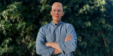 Barney Packer, a white man with blond hair, looking at the camera with his arms crossed. Behind him is green foliage.