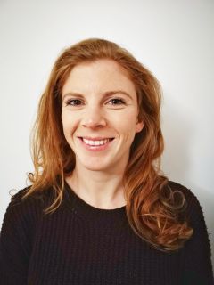 Lucy Scaramanga, a white woman with shoulder-length ginger hair, looks at the camera smiling. She wears a black jumper