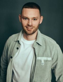 A portrait of James Hacking, a white man with short hair stubble, wearing a light jacket and white t-shirt