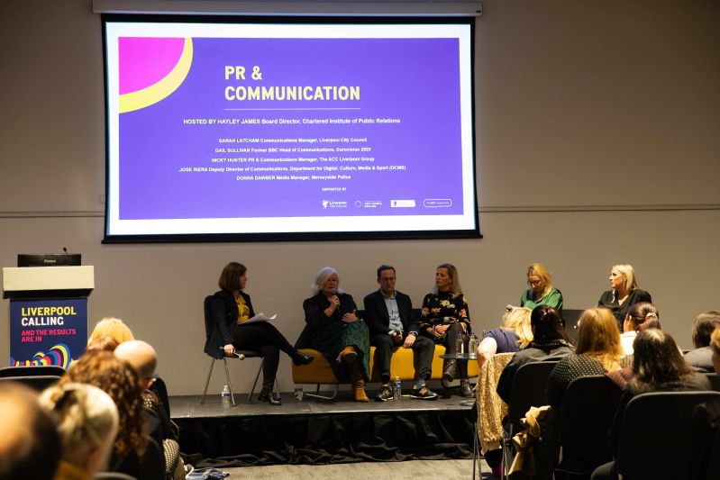Hayley James sat on stage with Donna Dawber, Jose Riera, Nicky Hunter, Gail Sullivan and Sarah Latcham. Above them is a screen which reads PR & Communication. A lecturn with Liverpool Calling is on the left. The backs of some audience members are visible.
