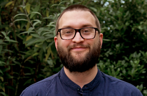 Phil Treagus-Evans, a white man with cropped brown hair, a bear, and glasses. Phil is photographed in front of a bush