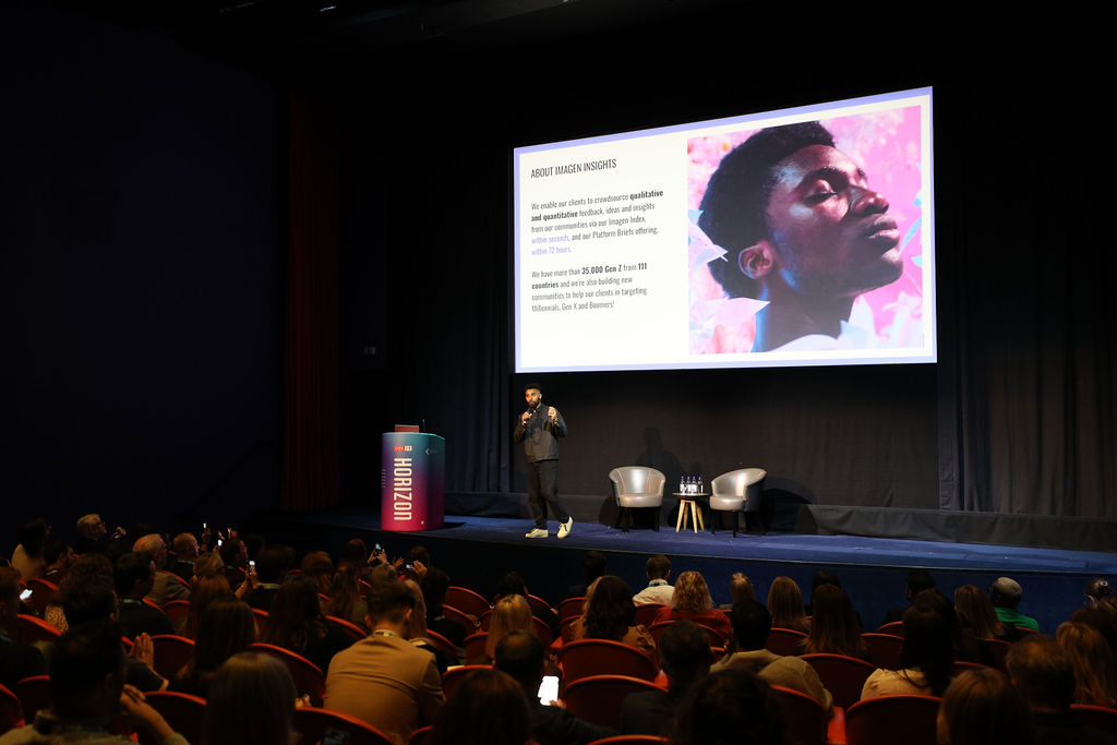 Jay Richards on stage at CIPR Horizon in front of an audience of dozens of people. He stands in front of a large screen displaying text and an image of the face of a black man