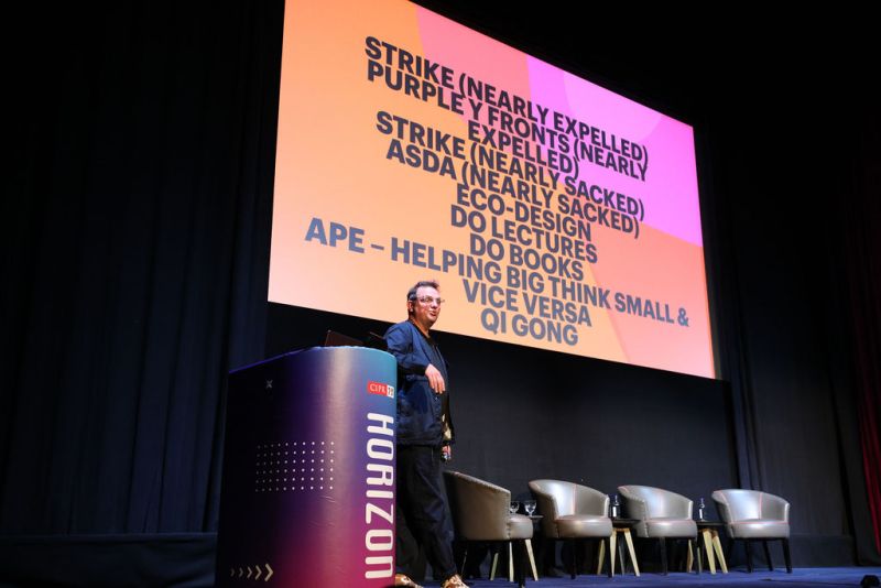 Mark Shayler, a white man wearing a navy jacket and trousers, on stage. To the left is a podium, to his right are four empty chairs. Above is a screen with his career information