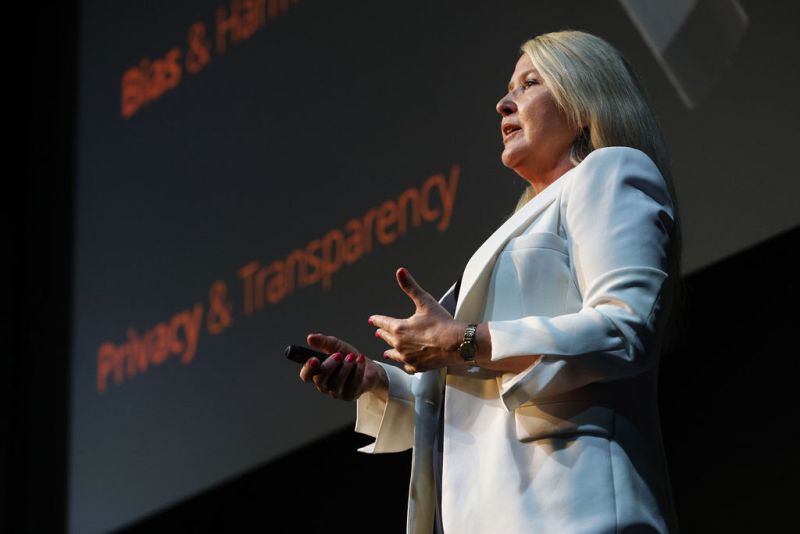 Katie King, a white woman with blonde hair, wears a white jacket. She holds a clicker in her right hand. She stands to the side of a large screen