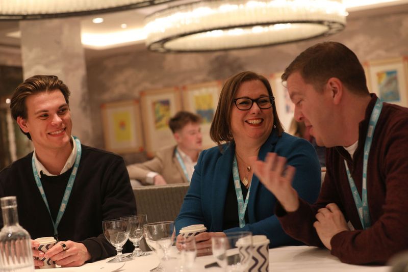 Three people sat at a table smiling and in conversation. In the centre is Rachel, a white woman with dark brown shoulder length hair, and Dan, a white man with short brown hair.