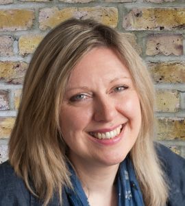 Hilary Berg smiling in front of a brick wall. Hilary is a white woman, with shoulder light brown hair 