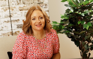 Louise Turner, a white woman with shoulder length red hair and wearing a pink patterned top, smiles at the camera. To her left is a green leafy plant. Behind is a brick wall