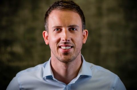 Stevie Brown, a white man with short brown hair smiles at the camera. He wears a blue shirt. The background is dark