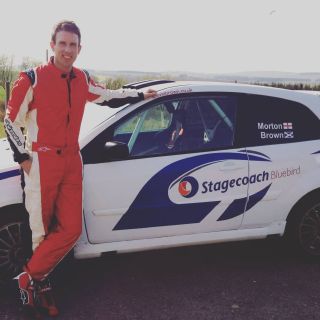 Stevie Brown, a white man wearing a red jumpsuit, stands next to a white car with blue Stagecoach logo on the side