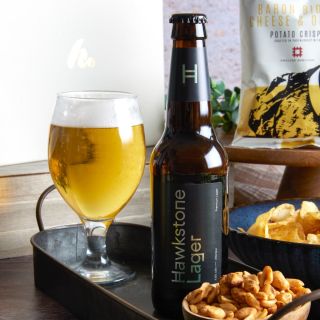 A glass of beer, a brown bottle of Hawkstone Lager, bowls of nuts and crips and a white bag of crisps