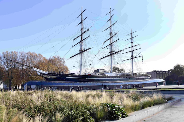 The Cutty Sark, a clipper ship with three masts, in dry dock on a bright day. There is planting in the foreground