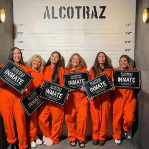 Eight white women wearing orange jumpsuits smile while in a line-up, posing with inmate signs under the venue's logo