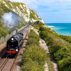A steam locomotive pulls carriages along a track cut into a hill alongside the sea. The sky is blue